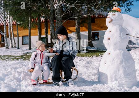 Mom füttert ein kleines Mädchen Haferbrei von einem Löffel, sitzt auf einem Schlitten neben einem Schneemann im Garten Stockfoto