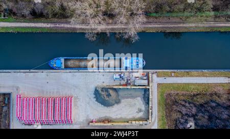 Dieses Bild zeigt eine Industrieszene aus der Vogelperspektive und zeigt ein großes blaues Frachtschiff, das ordentlich an einem Depot am Fluss angedockt ist. Das Schiff, gefüllt mit gerundeten Frachtcontainern, befindet sich parallel zum baumgesäumten Ufer, was einen starken Kontrast zum organisierten Chaos der daneben liegenden Reederei darstellt. Stapel von roten und weißen Frachtcontainern sind ordentlich in Reihen angeordnet, wodurch ein farbenfrohes Muster auf dem grauen Beton und den sandigen Materialien des Depots entsteht. Das ruhige Wasser reflektiert das sanfte Licht eines bewölkten Himmels, während die Umgebung von ruhiger Aktivität spricht Stockfoto