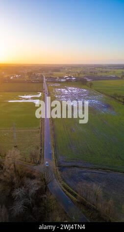 Dieses Luftbild fängt die ruhige Schönheit der Landschaft während des Sonnenuntergangs ein. Eine gerade Landstraße durchzieht die Landschaft und führt zum Horizont, wo das letzte Leuchten der Sonne den Himmel berührt. Die Felder auf beiden Seiten sind vom goldenen Licht der Sonne berührt, mit Landflächen, die die leuchtenden Farben des Himmels reflektieren. Die untergehende Sonne wirft lange Schatten und verleiht der Szene Tiefe und Kontrast. Dieser friedliche Moment verkörpert die weiten offenen Plätze und den ruhigen Abschluss eines Tages in ländlichen Umgebungen. Luftaufnahme der Country Road bei Sonnenuntergang mit leuchtenden Feldern. Hochwertige Fotos Stockfoto