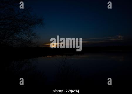 Dieses Foto fängt den dramatischen Kontrast des dunklen Abendhimmels mit den letzten glänzenden Strahlen der untergehenden Sonne über einem ruhigen See ein. Die Silhouetten der Bäume am See verleihen dem Bild einen zarten Rahmen und verstärken das Gefühl der Ruhe, das mit der nächtlichen Nacht einhergeht. Der Horizont wird von einem schmalen Lichtband beleuchtet, das vom stillen Wasser reflektiert wird, das als natürliche Leinwand für dieses ruhige Schauspiel dient. Die Szene verkörpert den täglichen Abschied des Lichts und lädt zur Reflexion und Introspektion ein. Silhouette bei Einbruch der Dunkelheit am Seeufer. Hochwertige Fotos Stockfoto