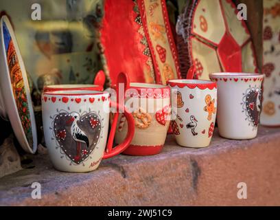 Riquewihr, Frankreich - 2. Juni 2023: Keramikbecher mit traditionellen elsässischen Motiven, Störche, Lebkuchenmann und Brezeln in einem Souvenirladen Stockfoto