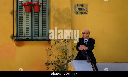 Menton, Frankreich - 26. Mai 2023: Glatzkopf in schwarzem Anzug und Krawatte mit Sonnenblume im Ohr sitzt auf einer Balustrade neben einem bunten traditionellen Haus Stockfoto