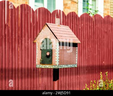 Briefkasten in Form eines kleinen Hauses auf dem Hintergrund eines roten Zauns Stockfoto