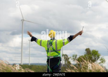Junger Ingenieur, der auf einem Feld für Windkraftanlagen arbeitet, trägt eine Sicherheitsweste. Heben Sie beide Hände, um sich nach Abschluss der Windkraftanlagen zu entspannen Stockfoto