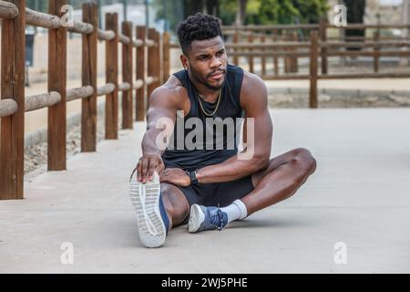 Der ganze Körper eines jungen starken afroamerikanischen Läufers sitzt auf dem Boden und dehnt die Beinmuskeln aus, während er vor dem Training die Zehen zieht Stockfoto