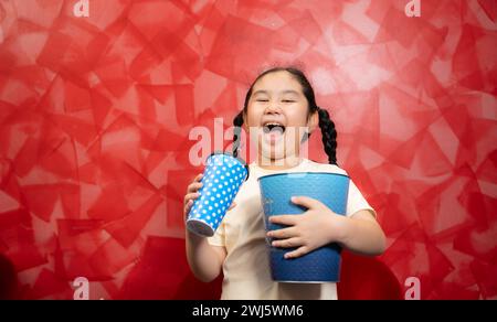 Asiatisches Kindermädchen, das Popcorn isst und Limonade auf roter Wand Hintergrund trinkt, Cinema Concept, Stockfoto