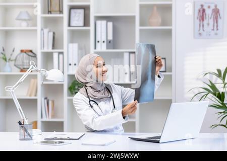 Lächelnde muslimische Ärztin im Hijab, die eine Röntgenuntersuchung in einem gut beleuchteten medizinischen Büro überprüft und die Professionalität und Vielfalt des Gesundheitswesens widerspiegelt. Stockfoto