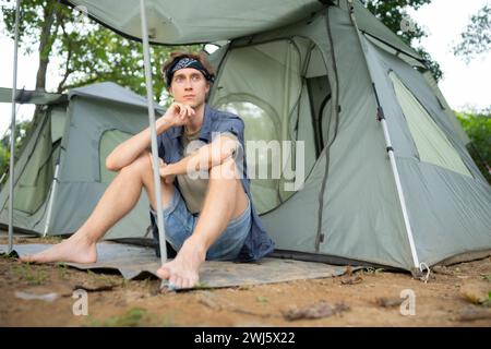 Junger Mann, der vor dem Zelt auf dem Campingplatz auf dem Land sitzt Stockfoto