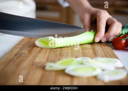 Nahaufnahme von Hand, Lauch mit Kochmesser auf einem Bambus-Schneidebrett, Tomaten im Hintergrund Stockfoto