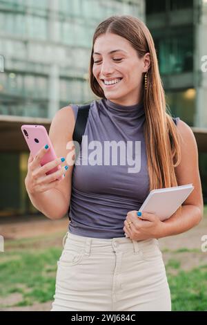 Vertikales Porträt einer glücklichen Teenagerin, die im Freien über eine Social-Media-App auf einem Smartphone lächelt und tippt. Eine junge Frau, die Spaß hat, Nachrichten auf einem Handy zu schreiben. Eine echte Dame, die ein Mobiltelefon benutzt. Hochwertige Fotos Stockfoto