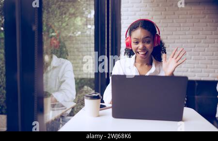 Porträt einer schönen jungen Frau, die Kopfhörer trägt und lächelt, während sie im Café ihren Laptop benutzt, und im Internet Freunde begrüßt Stockfoto