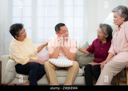 Senior asiatischer Mann, der an einer schweren Krankheit leidet, sitzt auf dem Sofa mit Freunden, die zu Besuch kommen, und ermutigt die Stockfoto