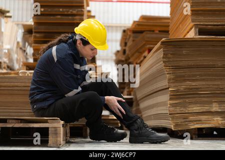 Lagerarbeiter, der auf dem Boden vor einem Stapel Pappkartons sitzt und Schmerzen im Bein hat, weil er auf Holzpaletten stößt. Stockfoto