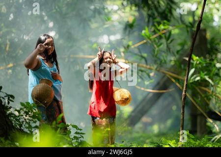 Zwei asiatische Mädchen mit traditioneller Kleidung stehen im Regenwald. Sie hatten Spaß miteinander zu spielen, bevor sie Opa halfen Stockfoto
