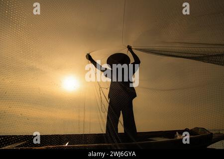 Silhouette eines Fischers bei Sonnenaufgang, an Bord eines Ruderbootes stehen und ein Netz werfen, um Fische nach Nahrung zu fangen Stockfoto