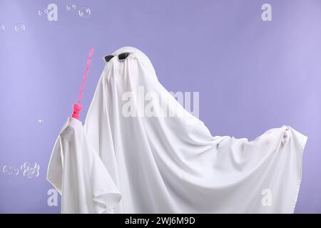 Lustiger Geist. Person in weißem Blatt und Sonnenbrille, die Seifenblasen auf violettem Hintergrund bläst Stockfoto