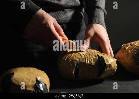 Schmuggel und Drogenhandel. Mann, der ein Paket mit Betäubungsmitteln öffnet, mit einem Cutter am dunklen Tisch, Nahaufnahme Stockfoto