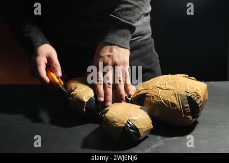 Schmuggel und Drogenhandel. Mann, der ein Paket mit Betäubungsmitteln öffnet, mit einem Cutter am dunklen Tisch, Nahaufnahme Stockfoto