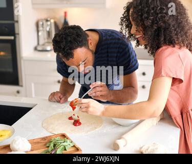 Ein Paar Zu Hause Mit Einem Mann Mit Down-Syndrom Und Einer Frau, Die In Der Küche Sauce Auf Pizza Legt Stockfoto