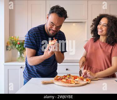 Ein Paar Zu Hause Mit Einem Mann Mit Down-Syndrom Und Einer Frau, Die Gemeinsam Hausgemachte Pizza In Der Küche Isst Stockfoto