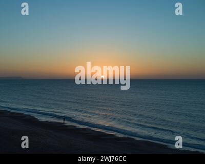 Ein einsamer Mann läuft an einem Wintermorgen bei Sonnenaufgang an der Flutlinie entlang. Es gibt keine Wolken am blauen und orangen Sonnenaufgangshimmel im Januar und das Meer ist ruhig. Stockfoto