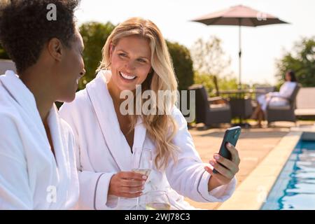 Zwei Reife Freundinnen Mit Handy In Bademänteln Draußen Am Pool Trinken Champagner Am Spa Day Stockfoto