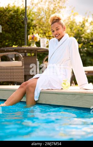 Porträt Einer Frau Im Bademantel, Die Im Freien Sitzt Und Mit Füßen Im Swimmingpool Sitzt, Mit Einem Getränk Am Spa Day Stockfoto