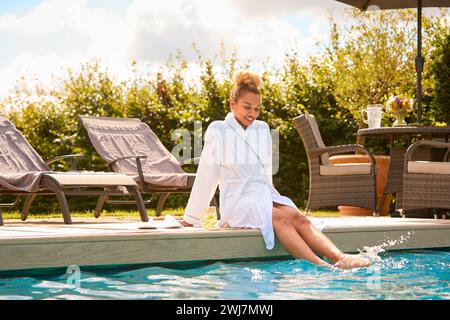 Frau Im Bademantel Sitzt Draußen Und Spritzt Füße Im Swimmingpool Am Spa Day Stockfoto