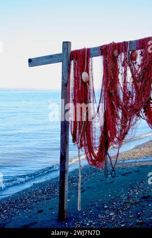 Rotes Fischernetz mit Bojen auf Holzpfosten am Strand, ruhiges Meer, klarer Himmel im Hintergrund. Stockfoto