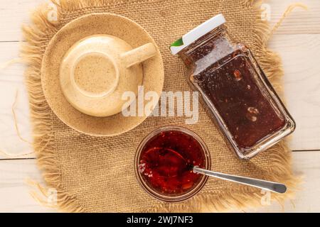 Erdbeermarmelade in Glasuntertasse mit Metalllöffel und Glasgefäß auf Juteserviette, Makro, Draufsicht. Stockfoto