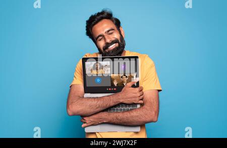 Ein zufriedener Mann mit Bart umarmt seinen Laptop liebevoll und zeigt ein virtuelles Meeting Stockfoto