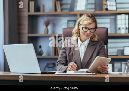 Lächelnde Geschäftsfrau, die im Büro arbeitet, an einem Schreibtisch sitzt, Notizen in einem Notebook macht und ein Tablet benutzt. Stockfoto