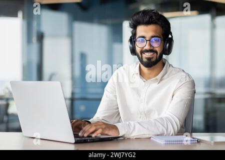 Porträt eines indischen Geschäftsmannes. Eine Tänzerin, die im Büro auf einem Laptop arbeitet, Kopfhörer trägt, sieht aus und lächelt in die Kamera. Stockfoto