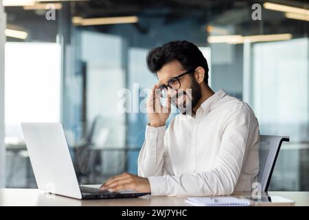 Junger indischer Mann Geschäftsmann und Büroangestellter, der unter schweren Kopfschmerzen und Spannungen leidet. Er sitzt an seinem Schreibtisch und hält seinen Kopf mit der Hand, zuckend vom Druck. Stockfoto