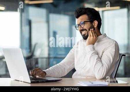 Zahnschmerzen bei einem jungen indischen Geschäftsmann. Am Arbeitsplatz im Büro am Tisch sitzen und eine Hand an die Wange halten, eine Massage durchführen, an Entzündungen leiden. Stockfoto