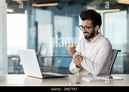 Ein indischer junger Mann leidet unter Schmerzen im Arm. Er sitzt im Büro am Schreibtisch und hält seine Hand am Handgelenk, gibt eine Massage. Stockfoto