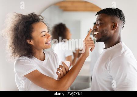 Verspielte Black Millennial Lady, die drinnen eine Feuchtigkeitspflege auf die Nase des Mannes aufträgt Stockfoto