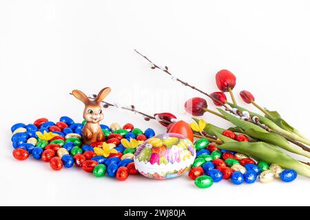 Eine Sammlung von bunten Schokoladeneiern und einem Osterhasen neben Tulpen und Palmkatzen, weißer Hintergrund Stockfoto