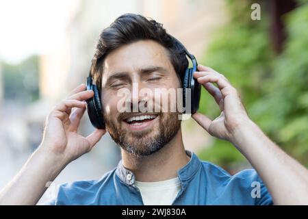 Lächelnder bärtiger Mann in lässiger Kleidung, der fröhlich Musik über Kopfhörer hört, während er abends in einer urbanen Umgebung spaziert. Stockfoto