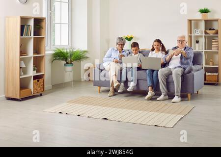 Glückliche Großeltern mit zwei Kindern, die zusammen mit Laptops und Mobiltelefonen auf dem Sofa sitzen. Stockfoto