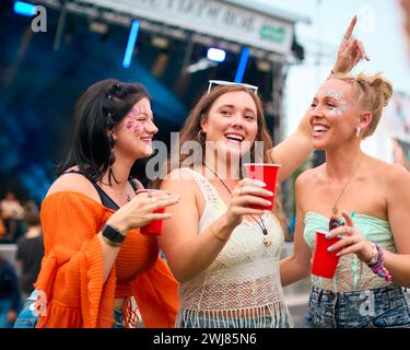 Drei Weibliche Freunde Mit Glitzer, Die Beim Summer Music Festival Spaß Haben Und Getränke Halten Stockfoto