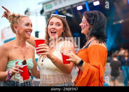 Drei Weibliche Freunde Mit Glitzer, Die Beim Summer Music Festival Spaß Haben Und Getränke Halten Stockfoto