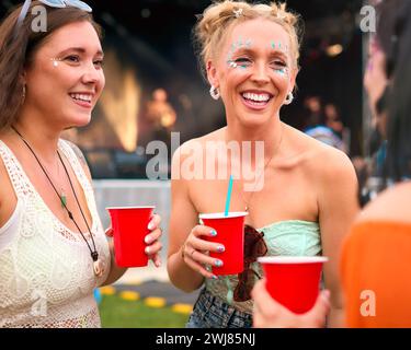 Drei Weibliche Freunde Mit Glitzer, Die Beim Summer Music Festival Spaß Haben Und Getränke Halten Stockfoto