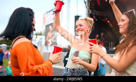 Drei Weibliche Freunde Mit Glitzer, Die Beim Summer Music Festival Spaß Haben Und Getränke Halten Stockfoto