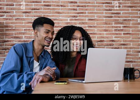 Ein vielseitiges Paar zu Hause, das online mit einem Laptop arbeitet. Lächelnder Mann und Frau, junger Erwachsener, der zusammen mit einem Laptop sitzt, während er mit einem Becher sitzt. Stockfoto