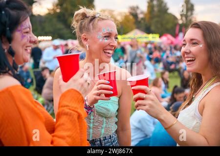 Drei Weibliche Freunde Mit Glitzer, Die Beim Summer Music Festival Spaß Haben Und Getränke Halten Stockfoto