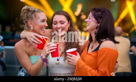 Drei Weibliche Freunde Mit Glitzer, Die Beim Summer Music Festival Spaß Haben Und Getränke Halten Stockfoto