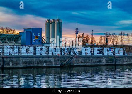 Blick vom Europakai auf eine bemalte Wand mit dem Wort Nürnberg an einem Gewerbebetrieb im Nürnberger Hafen. Im Hintergrund der Nürnberger Fernmeldeturm im Sonnenuntergang. Nürnberg Bayern Deutschland *** Blick von Europakai auf eine bemalte Wand mit dem Wort Nürnberg auf ein Handelsunternehmen im Hafen von Nürnberg im Hintergrund der Nürnberger Telekommunikationsturm bei Sonnenuntergang Nürnberg Bayern Deutschland 20240213-6V2A2285-Bearbeitet Stockfoto