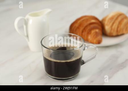 Aromatischer Kaffee in Glasbecher, Krug und frische Croissants auf weißem Marmortisch, Nahaufnahme Stockfoto