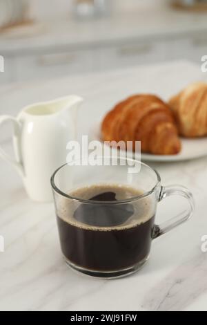 Aromatischer Kaffee in Glasbecher, Krug und frische Croissants auf weißem Marmortisch Stockfoto